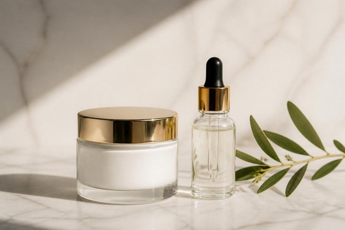 White cream jar with gold lid next to a serum bottle on marble